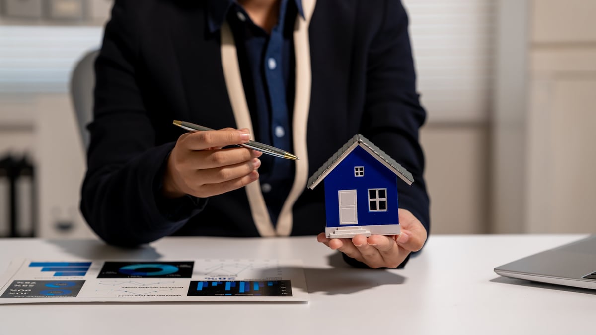 A person holding a model house beside property and finance charts